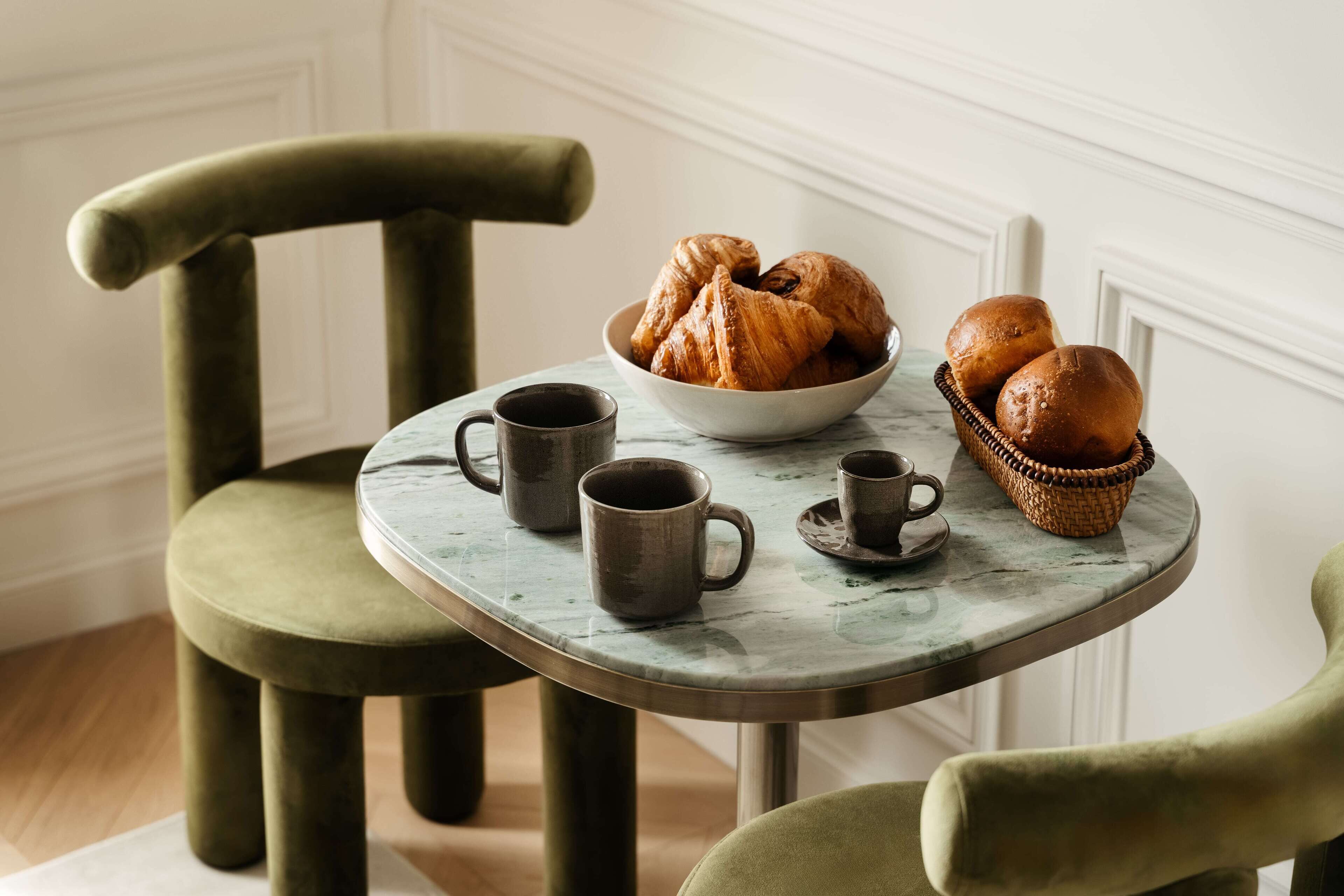 A dining table with a bowl of croissants, two coffee cups, and a basket of bread.