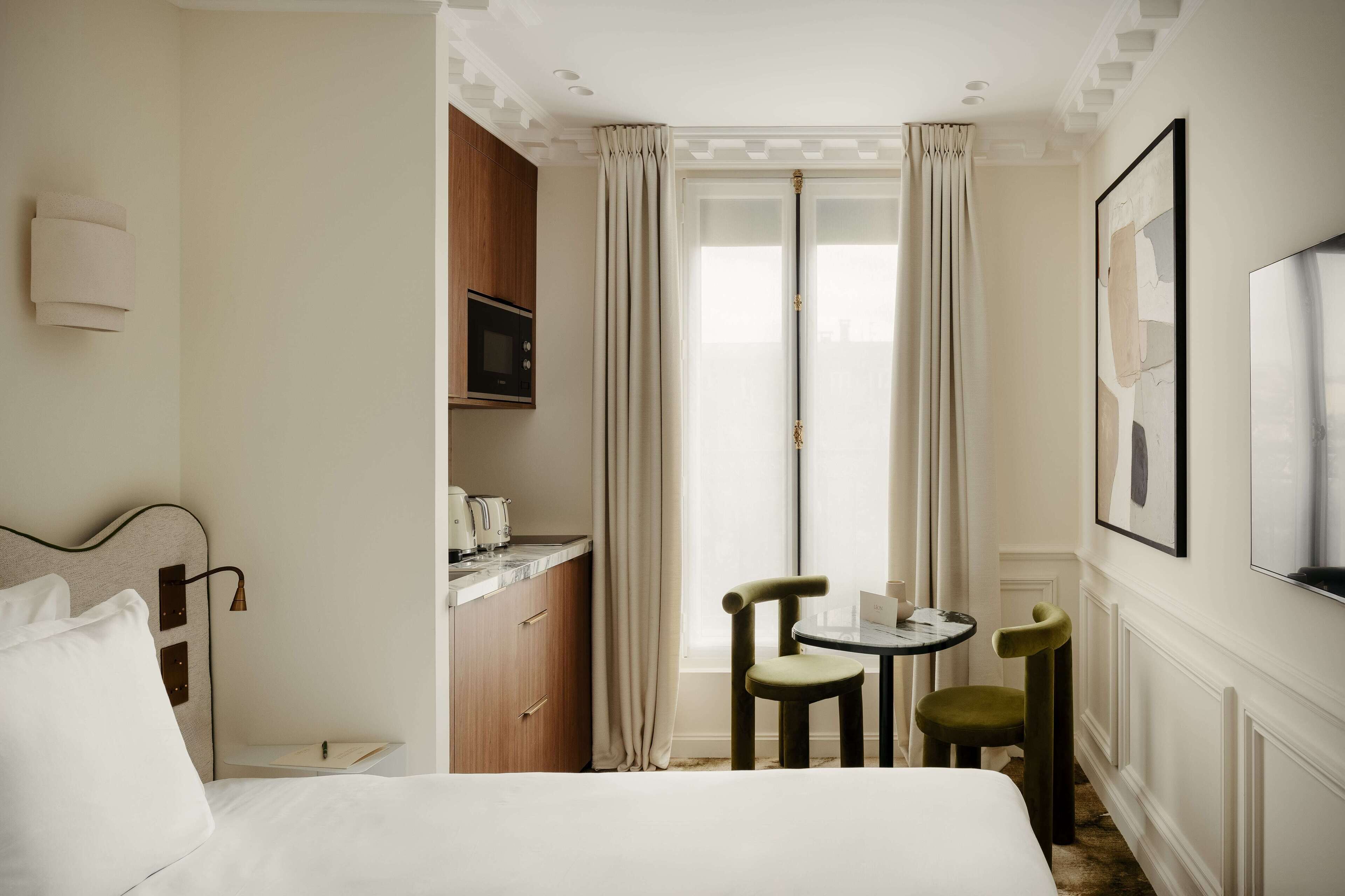 Una camera d'albergo con un letto, un angolo cottura, un'area pranzo con due sedie e un'ampia finestra con tende.