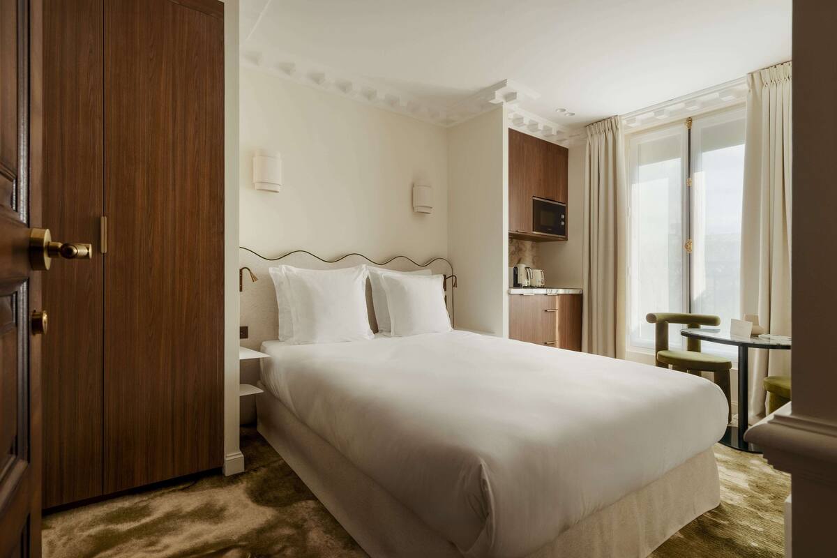 A hotel room with a large bed, a wooden wardrobe, a small table with a chair, and a TV.