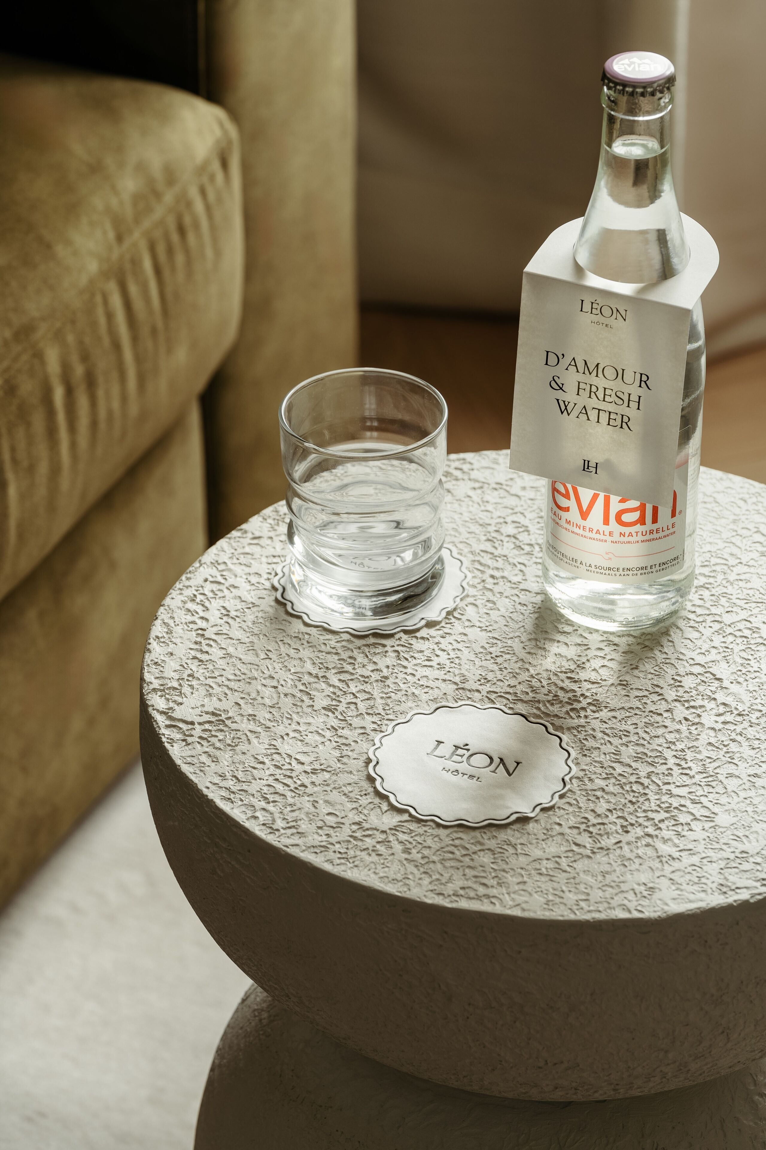 A bottle of Evian water and a glass on a textured table with a nameplate reading "LÉON HOTEL".