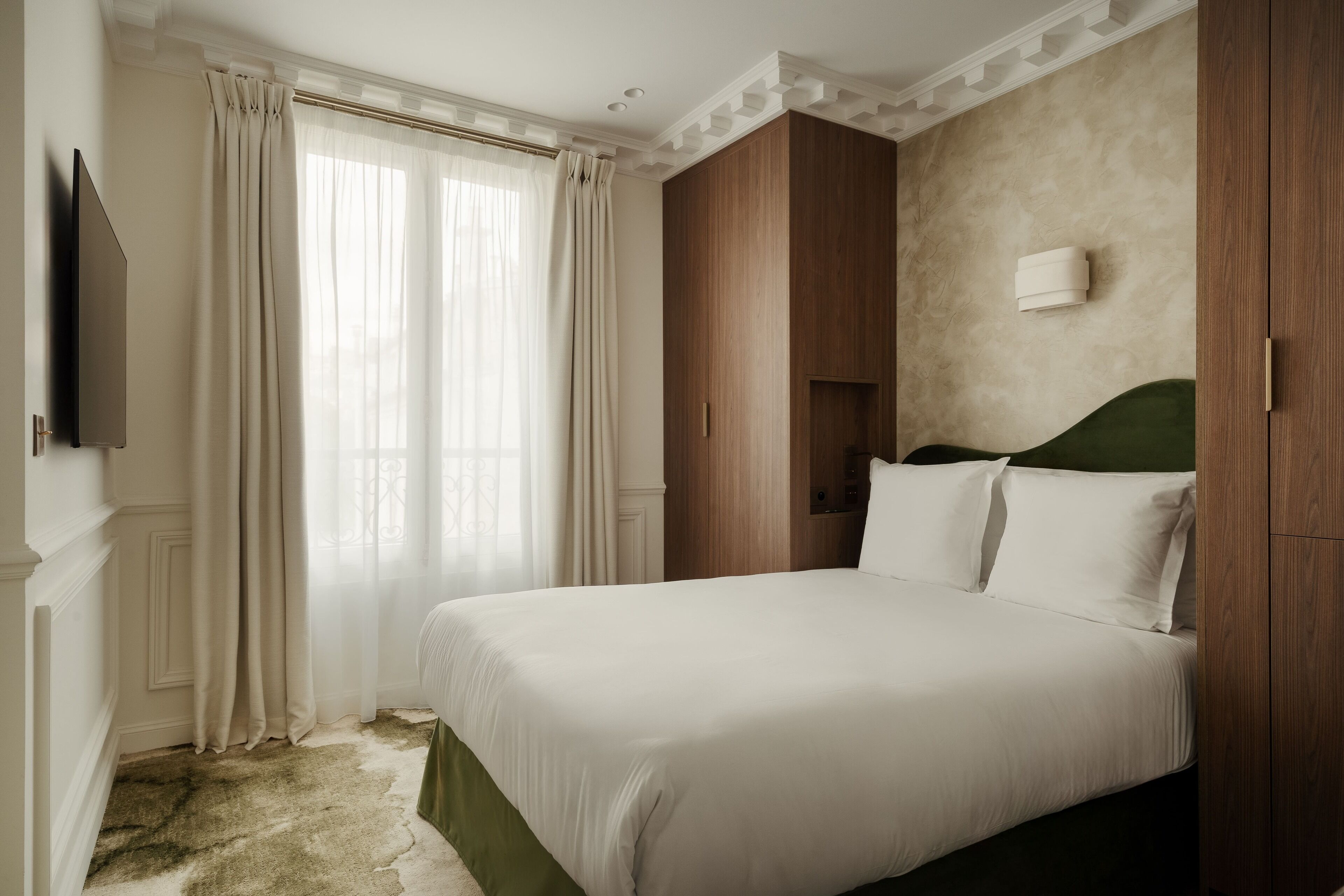 A hotel room with a large bed, wooden wardrobe, a flat-screen TV, and a window with curtains.