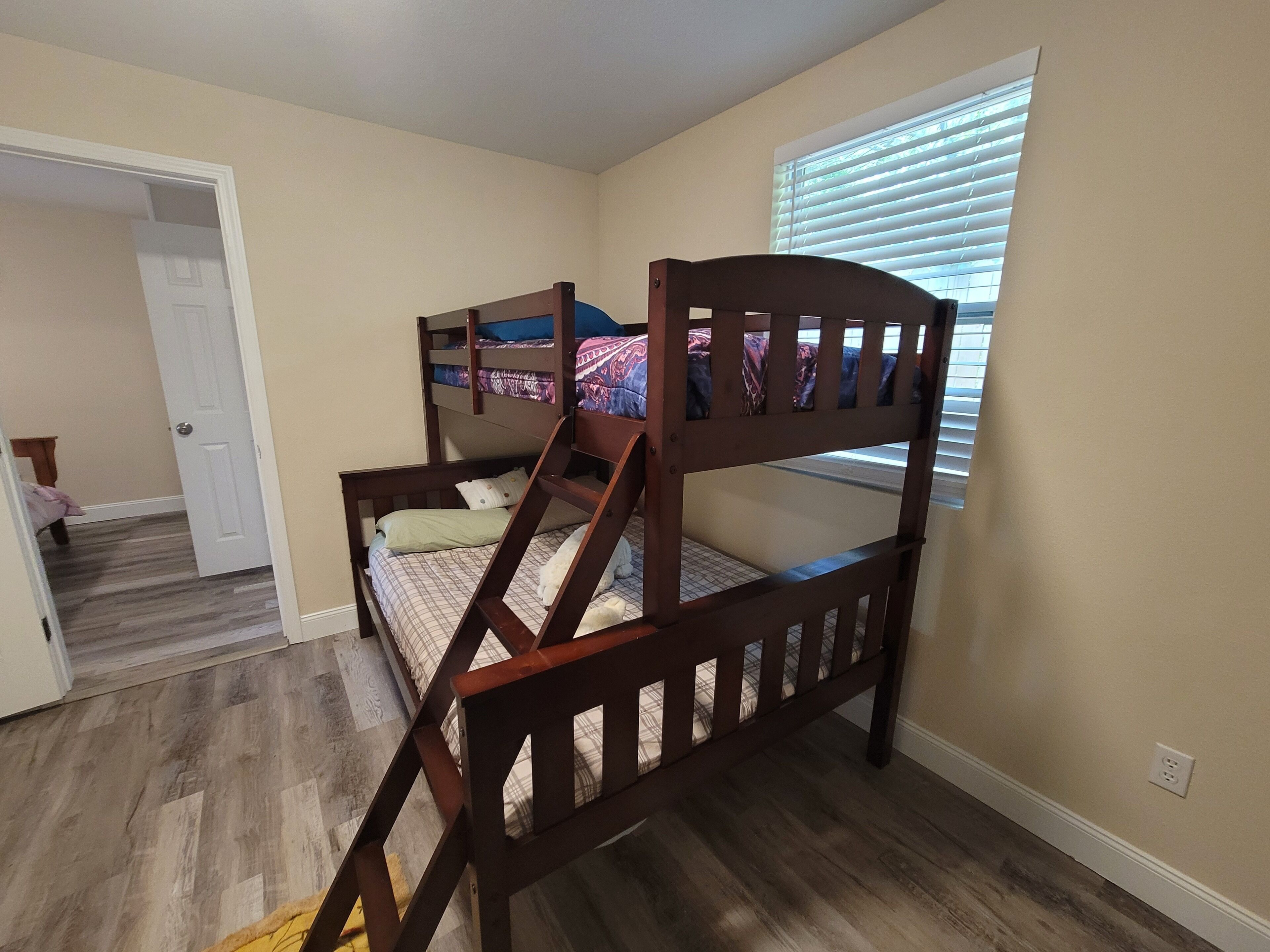 A bedroom with a bunk bed, wooden flooring, a door leading to another room, and a window with blinds.