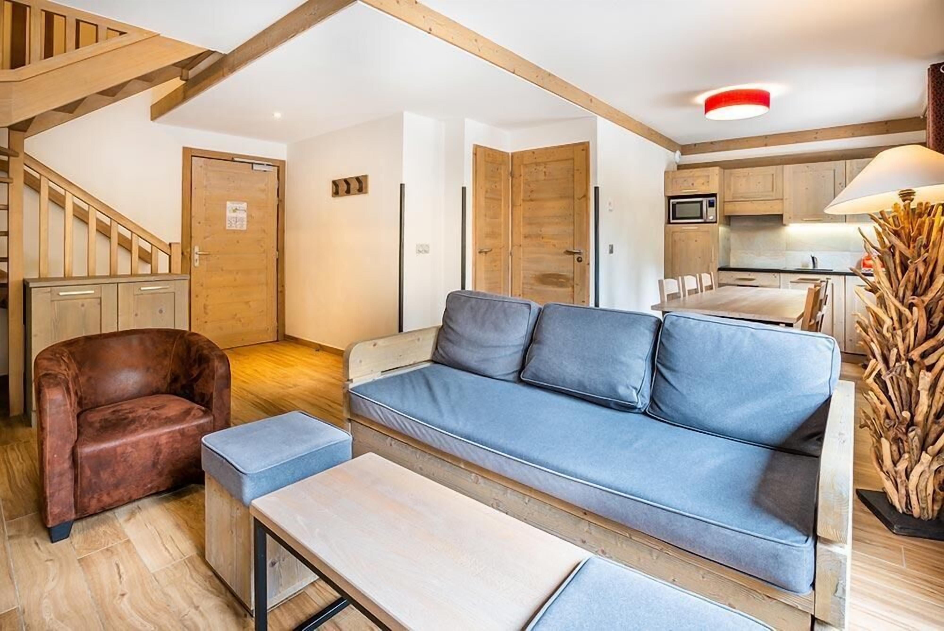 Un salon avec un canapé bleu, un fauteuil en cuir marron, une table basse en bois et un coin cuisine doté de meubles en bois et d’un four à micro-ondes.
