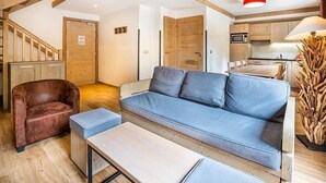 Una sala de estar con un sofá azul, un sillón de cuero marrón, una mesa de centro de madera y una zona de cocina con armarios de madera y un microondas.