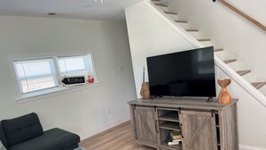 A modern living room with a grey sofa, a wooden entertainment unit with a flat-screen TV, a wicker basket, and a staircase.