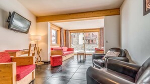 Un salon chaleureux, doté de meubles en bois, d’un téléviseur et offrant une vue sur un bâtiment en pierre grâce à de grandes fenêtres.