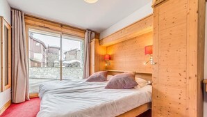 A bedroom with a large bed, wooden headboard, two wall-mounted lamps, and a view of buildings through a large window. - Wifi, 2nd floor, terrace, tv, ski locker, 59m², Tignes (Tignes)