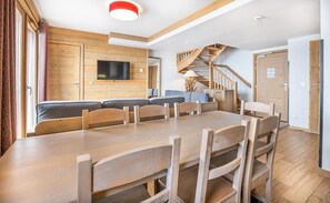 Een moderne woonkamer met een houten eettafel, stoelen, een bank en een aan de muur bevestigde televisie.