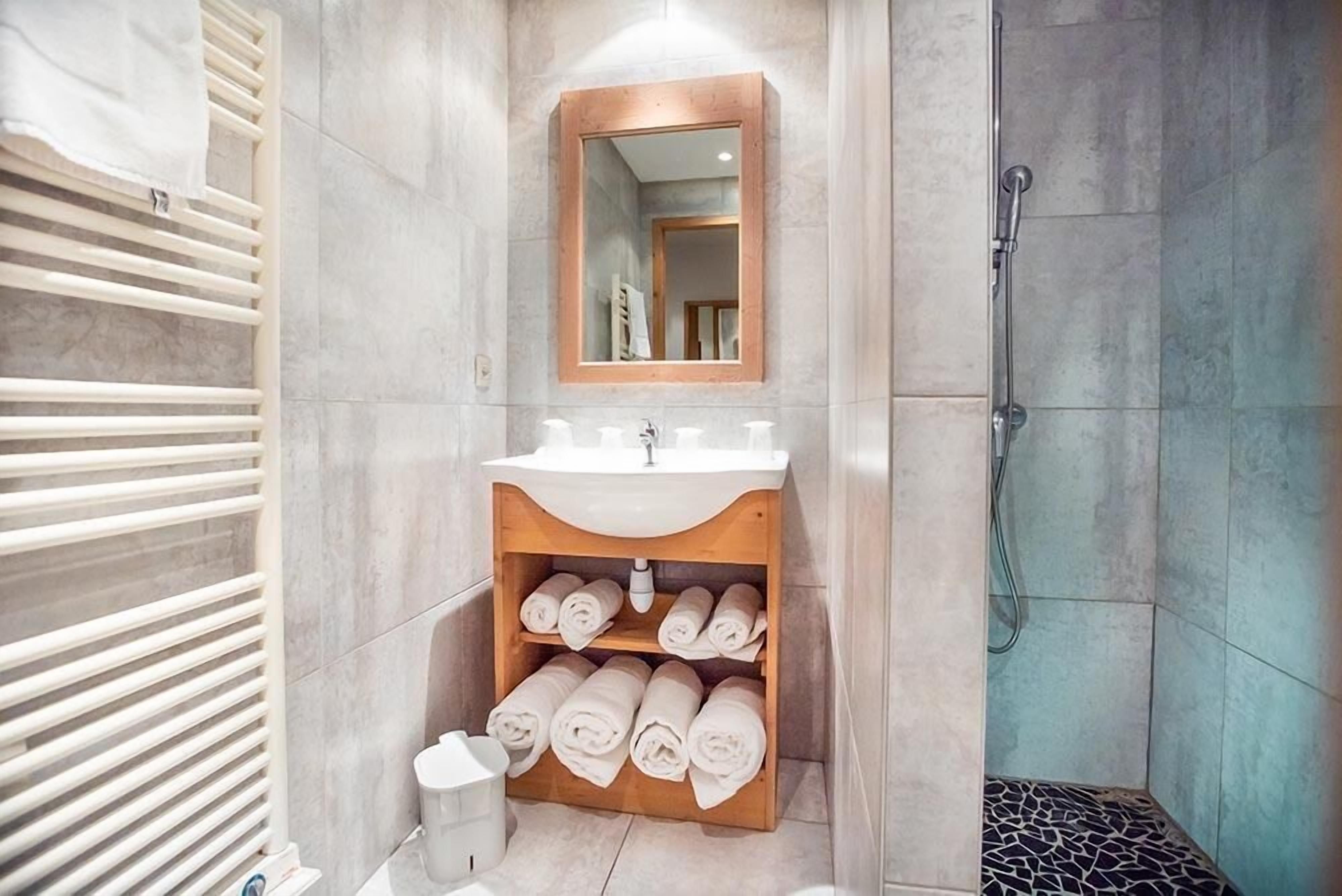 A bathroom with a wooden vanity, a mirror, a shower, and towels neatly rolled up.