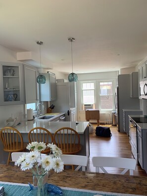 A modern kitchen with white cabinets, a central island with a sink, and a dining area with wooden chairs and a vase of flowers. - Beautifully home in Newport RI. Five star ratings in all previous rentals. (Newport)