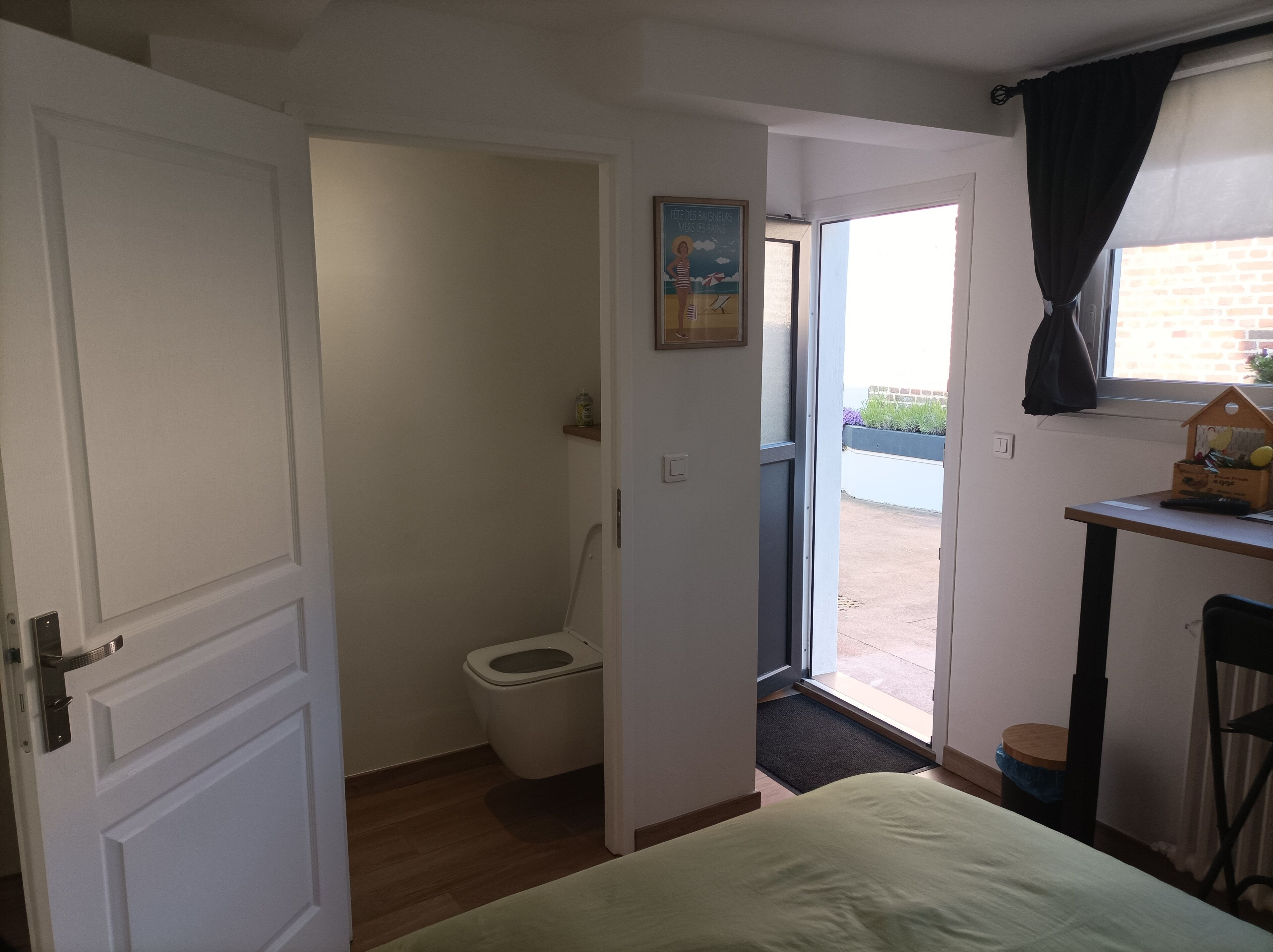 A room with a bed, a toilet, a small table with a lamp, and a window with curtains.