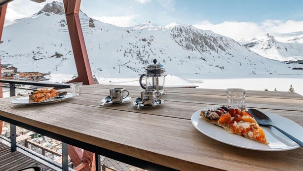 A wooden table with a plate of pizza, a coffee pot, and a glass of water, overlooking a snowy mountain landscape. - 200m from resort center, 3rd floor, view lake, balcony, ski locker, 75m², Tignes (Tignes)