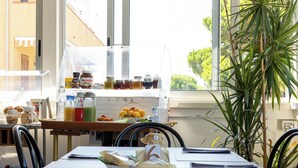 Desayuno con diversos alimentos y condimentos sobre una mesa, una planta y vistas por la ventana.