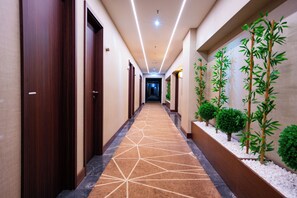 Un pasillo de hotel con una alfombra de diseño geométrico, puertas de madera y una fila de plantas verdes a la derecha.