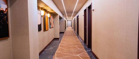 A hotel corridor with a patterned carpet, wall-mounted lights, and doors on both sides.
