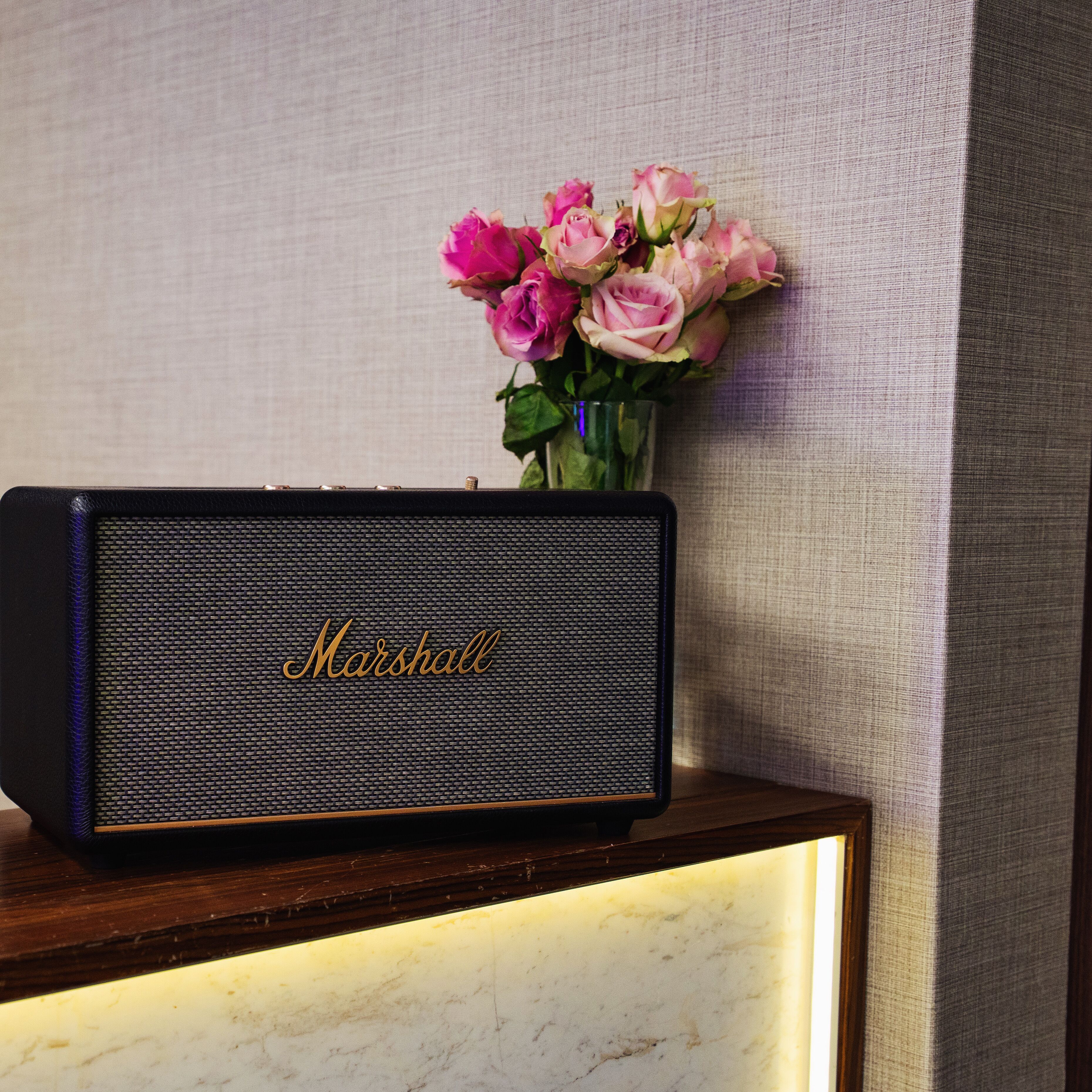 Un altavoz Marshall sobre una mesa de madera con un jarrón de flores rosas al fondo.