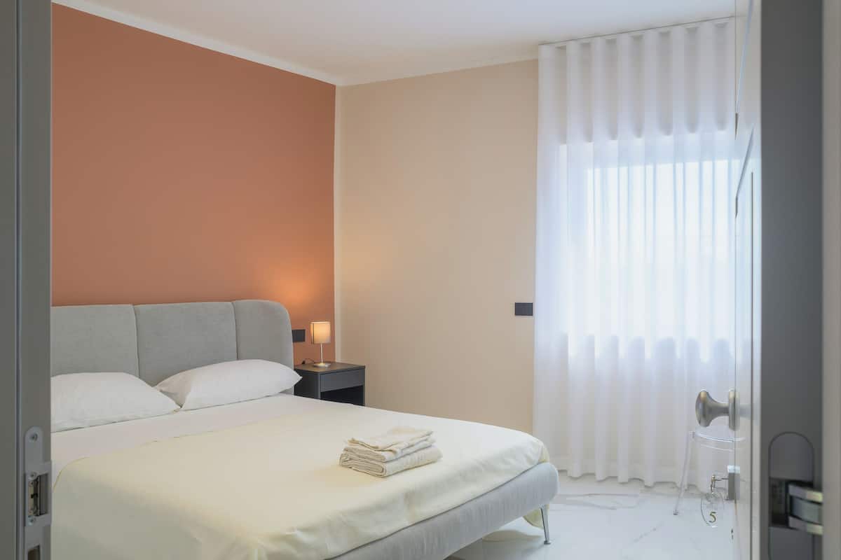 A neatly made bed with a white comforter and pillows, a bedside table with a lamp, and a window with sheer curtains.