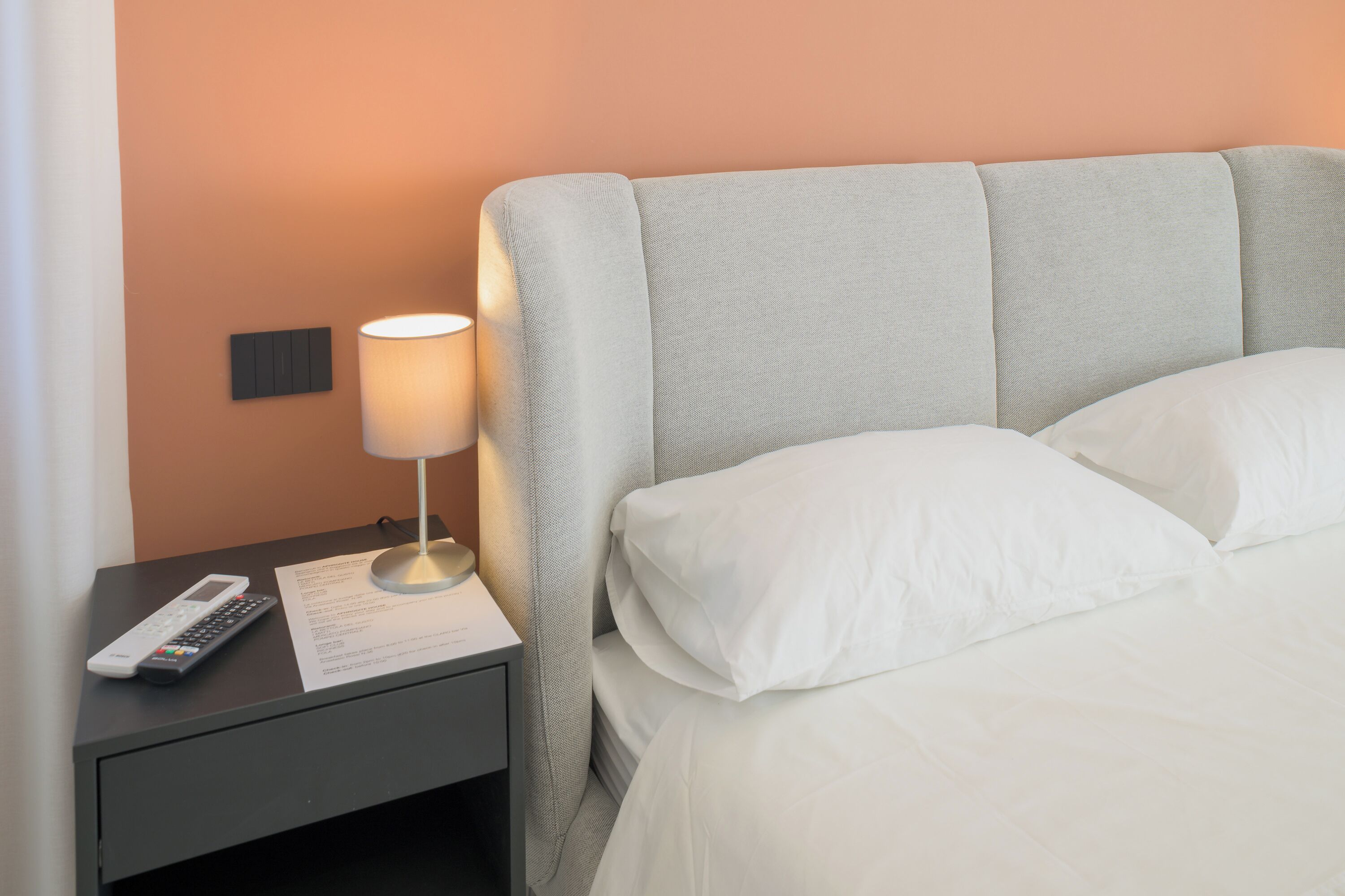 A neatly made bed with white linens, a bedside lamp, a remote control, and a notepad on a nightstand.