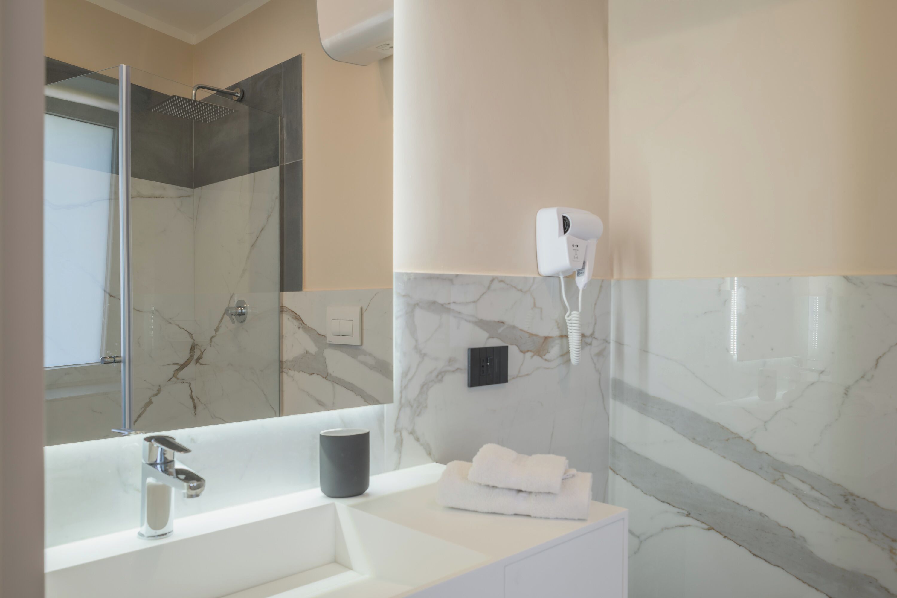A modern bathroom with a large mirror, a white sink, a handheld showerhead, and a hairdryer mounted on the wall.