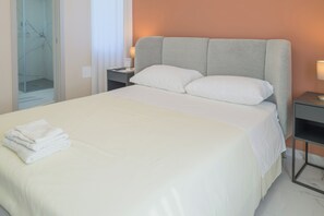 A hotel room with a neatly made bed, a nightstand with a lamp, and a glass shower enclosure. - Aphrodite House (Pompei)