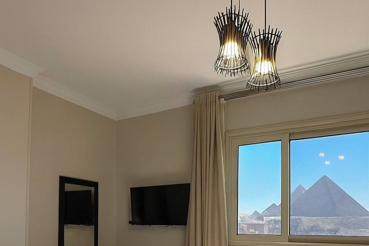 A hotel room with a bed, a television, a window with a view of pyramids, and a chandelier.