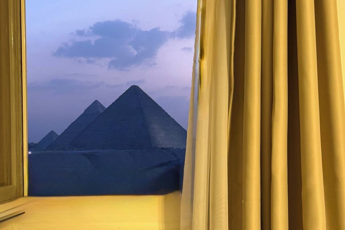 A window with curtains, revealing a view of pyramids under a clear sky.