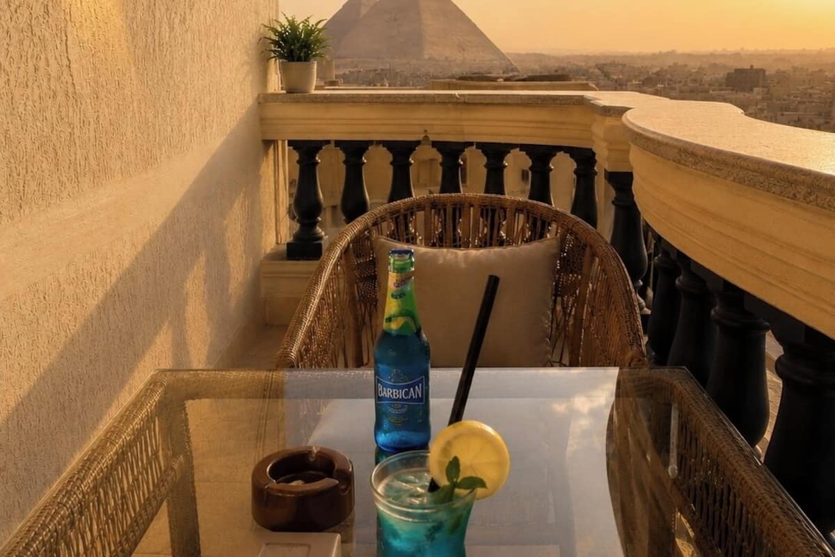 A balcony with a view of the pyramids, a table with a drink, and wicker chairs.