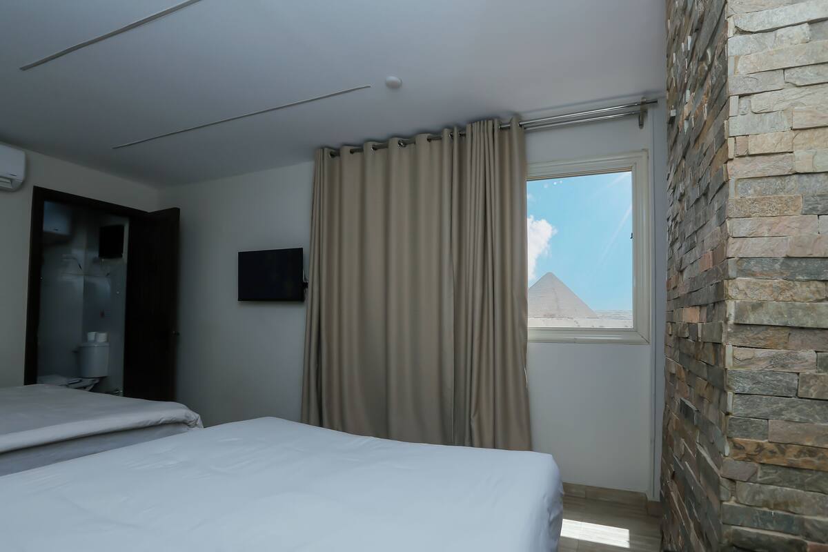 A hotel room with two beds, a stone accent wall, a TV mounted on the wall, and a window with a view of a pyramid.