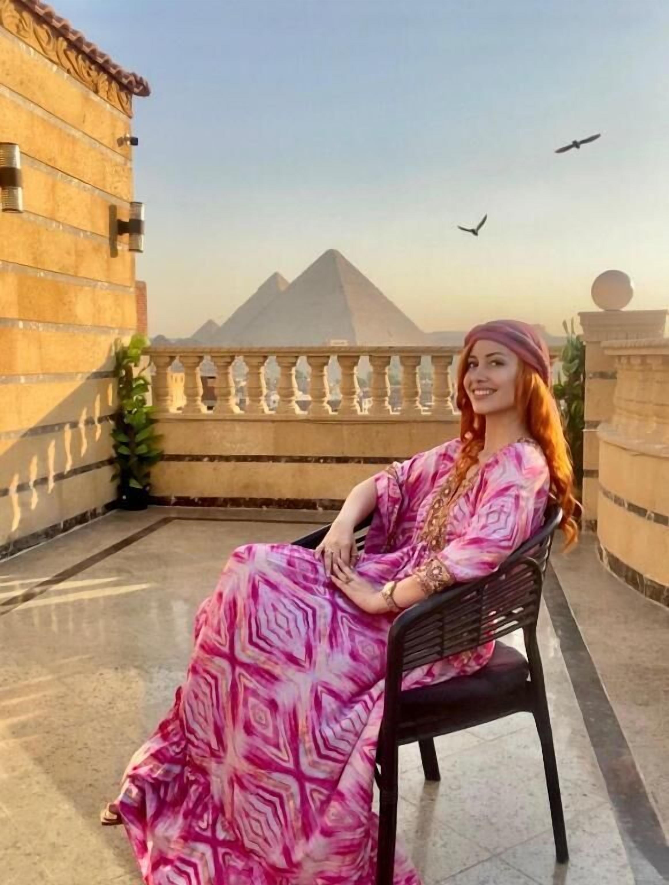 A person seated on a balcony with a view of a pyramid in the background.