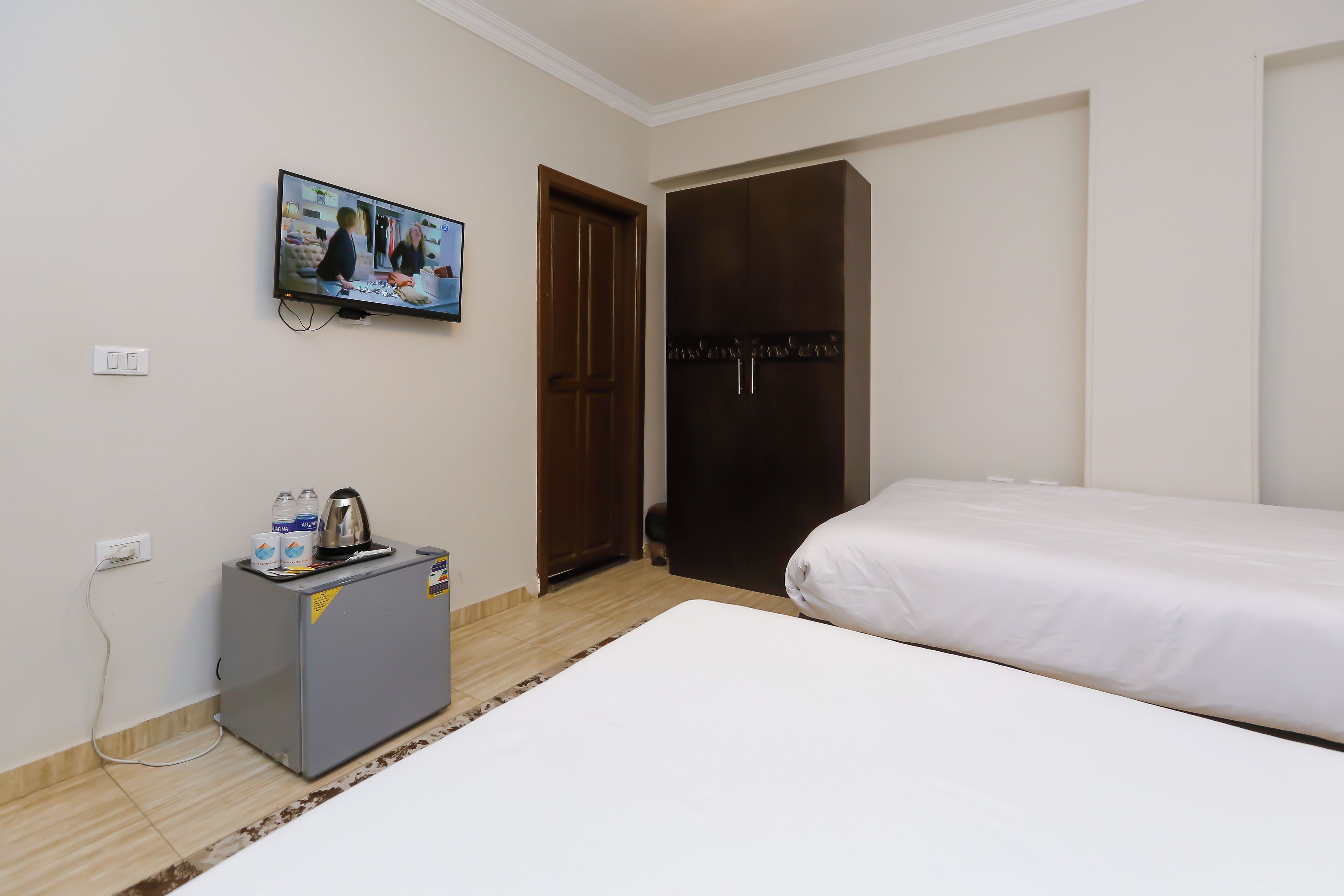 A hotel room with a bed, a TV mounted on the wall, a wardrobe, and a small refrigerator with beverages and a kettle.