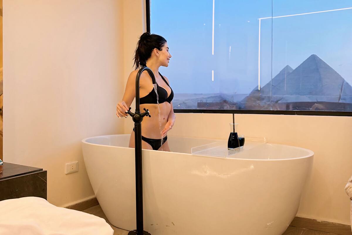A woman standing in a bathtub with a window view of pyramids.