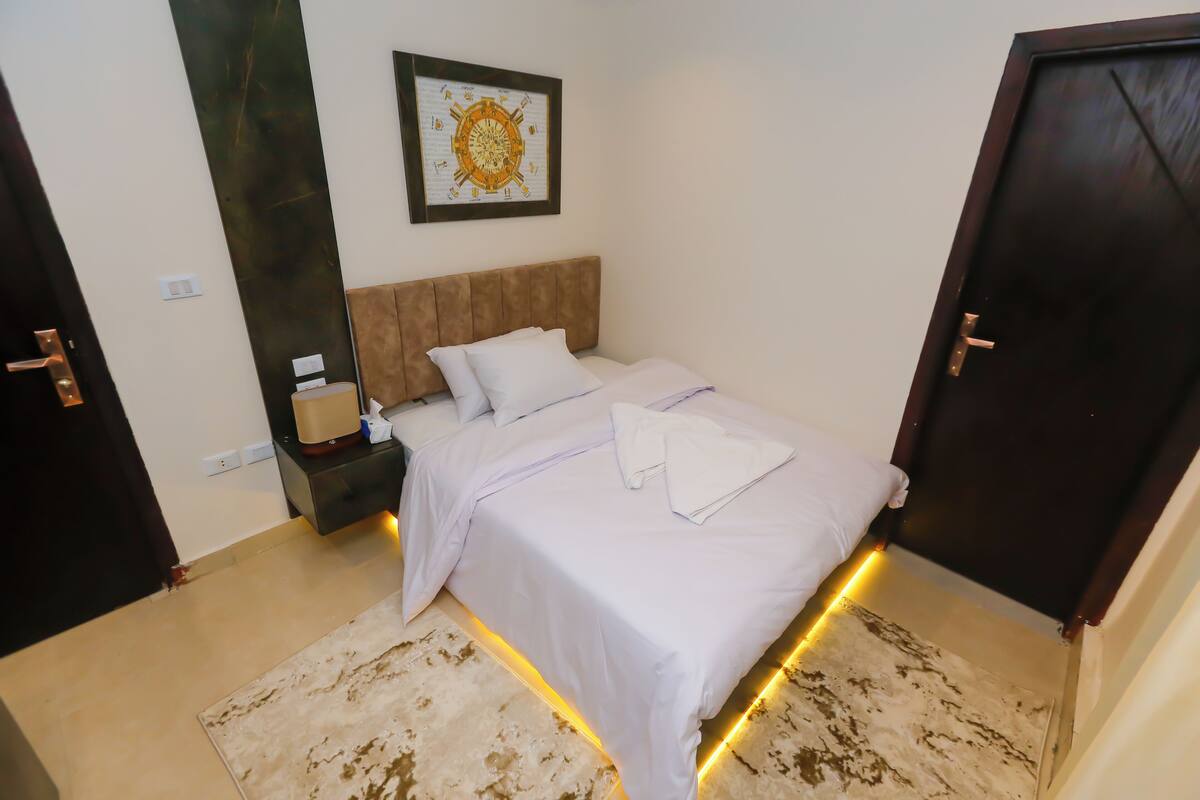 A neatly made bed with white linens, a bedside table with a lamp, and a wall-mounted artwork.