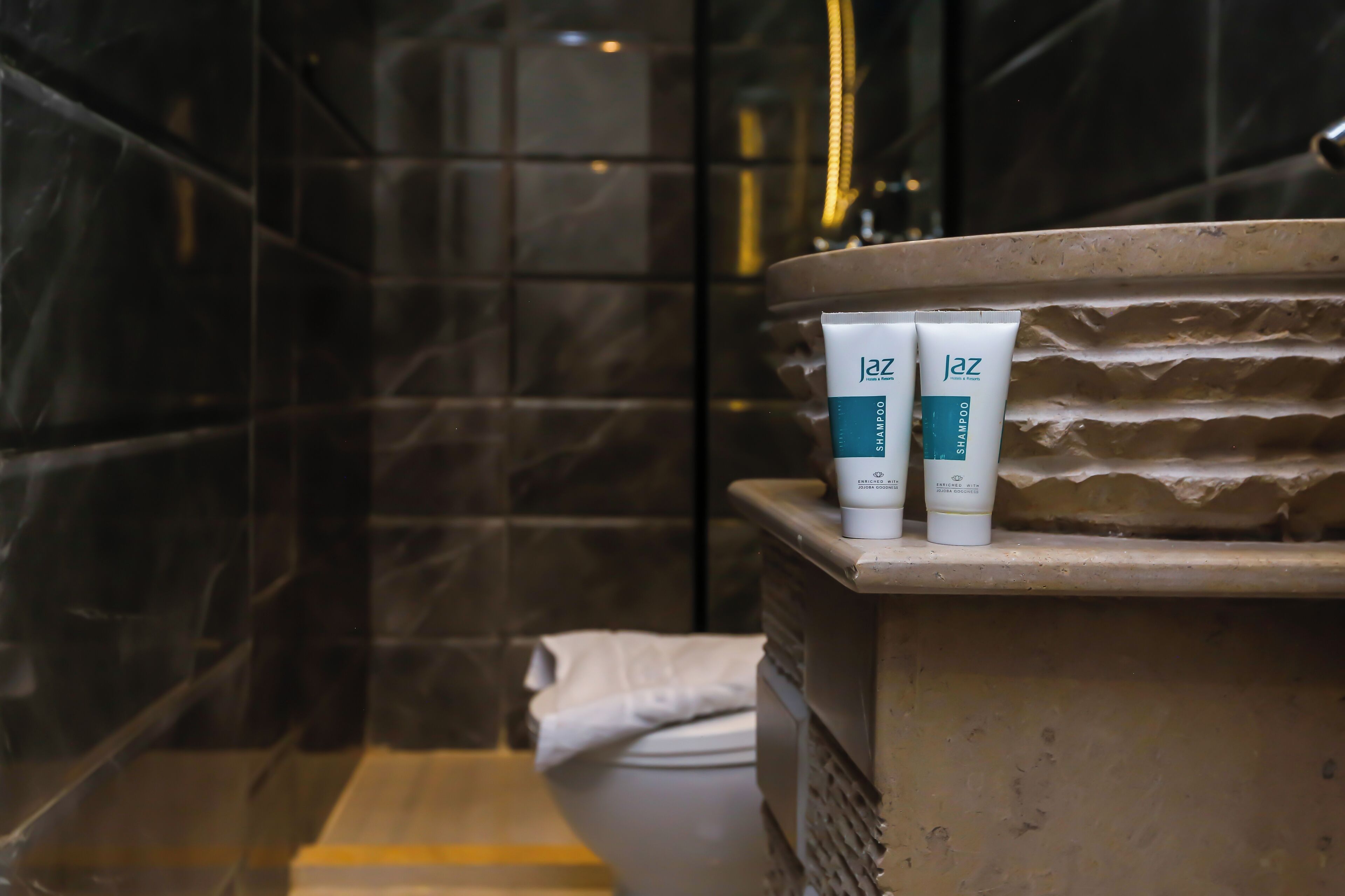 A bathroom with a marble wall, a bathtub, and two tubes of 'jaz' shampoo on the edge.