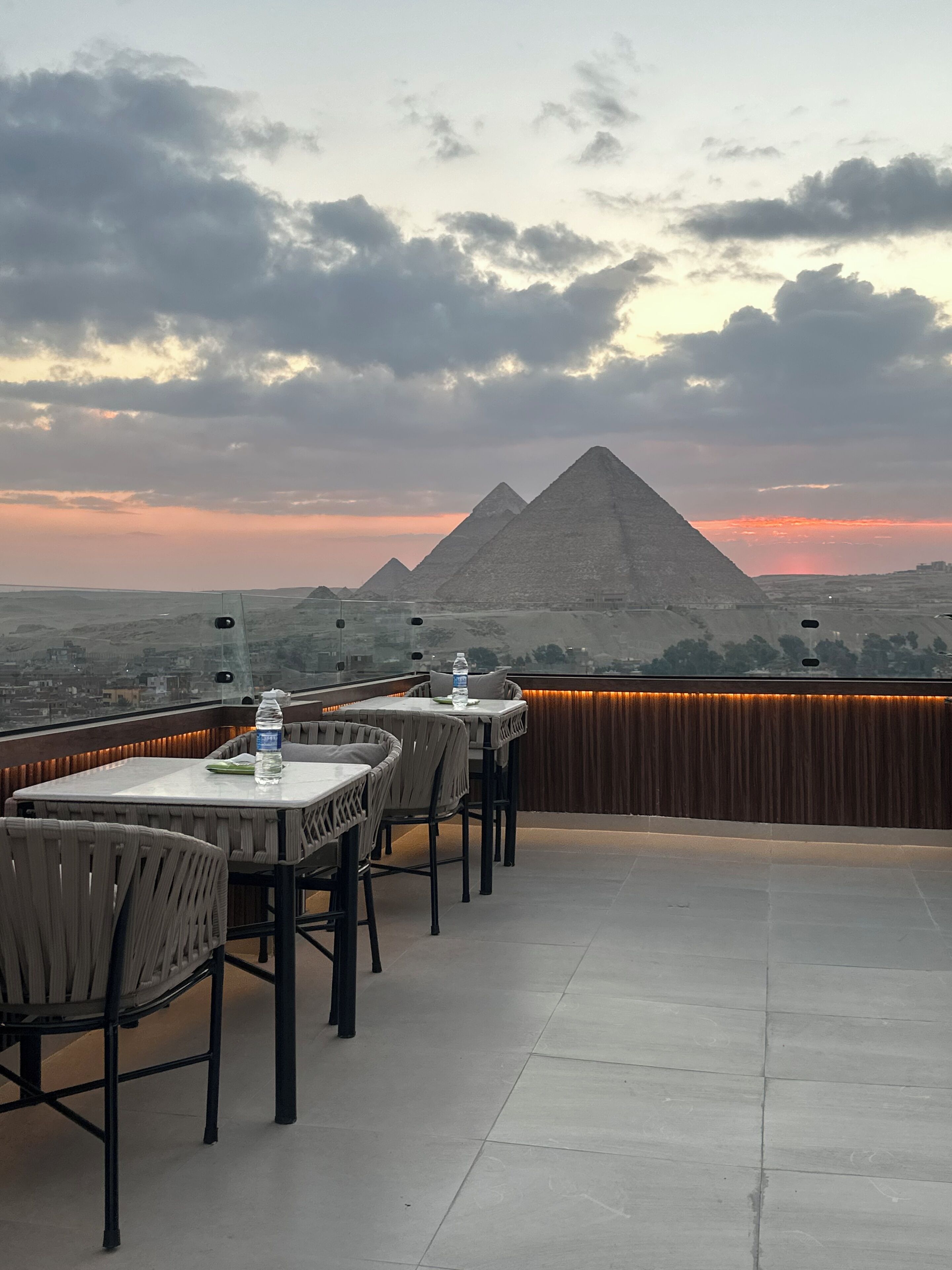 A rooftop terrace with tables and chairs, overlooking a desert landscape with pyramids in the distance.