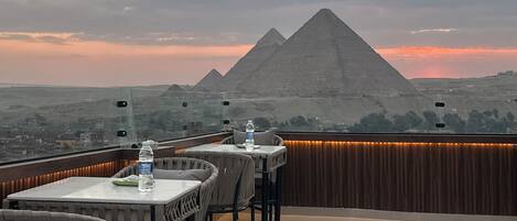 A rooftop terrace with tables and chairs, overlooking a desert landscape with pyramids in the distance.