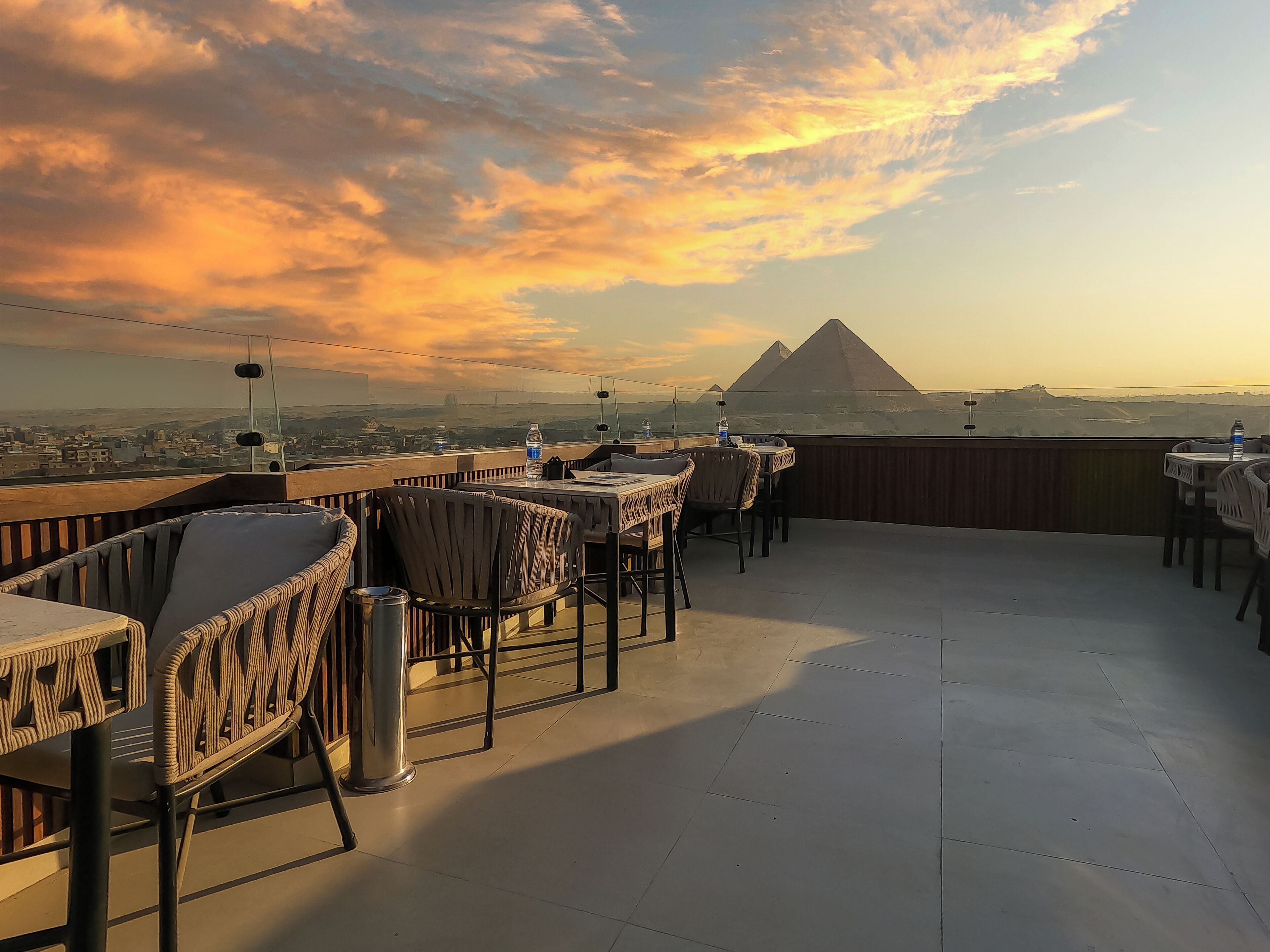 A rooftop terrace with tables and chairs, a view of a pyramid, and a sunset sky.