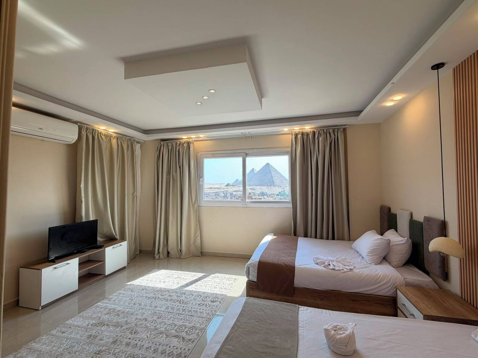A hotel room with a bed, a TV, a window with a view of a pyramid, and an air conditioning unit.