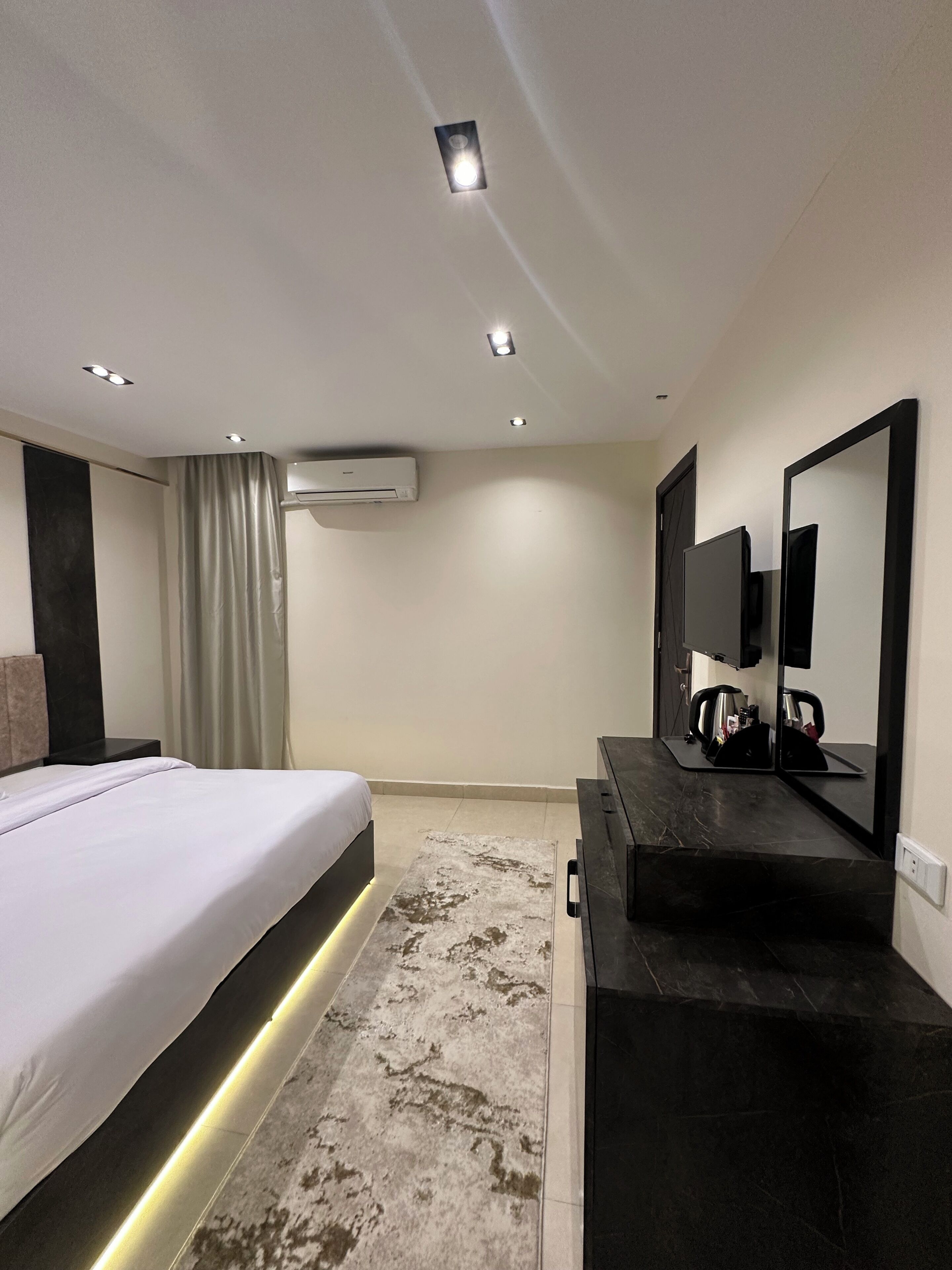 A modern hotel room with a bed, a dark wooden dresser, a large mirror, and an air conditioning unit on the wall.