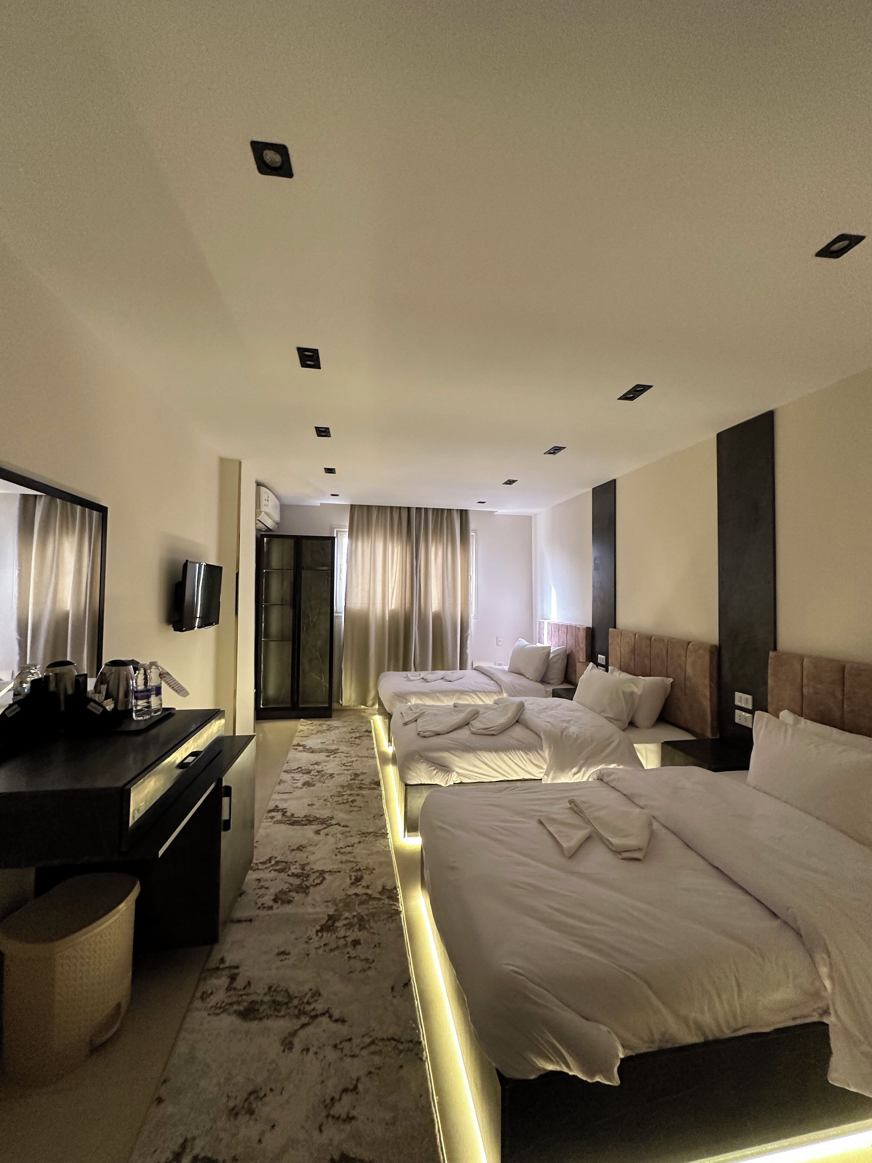 A hotel room with three beds, a desk with a flat-screen TV, and a large window with curtains.