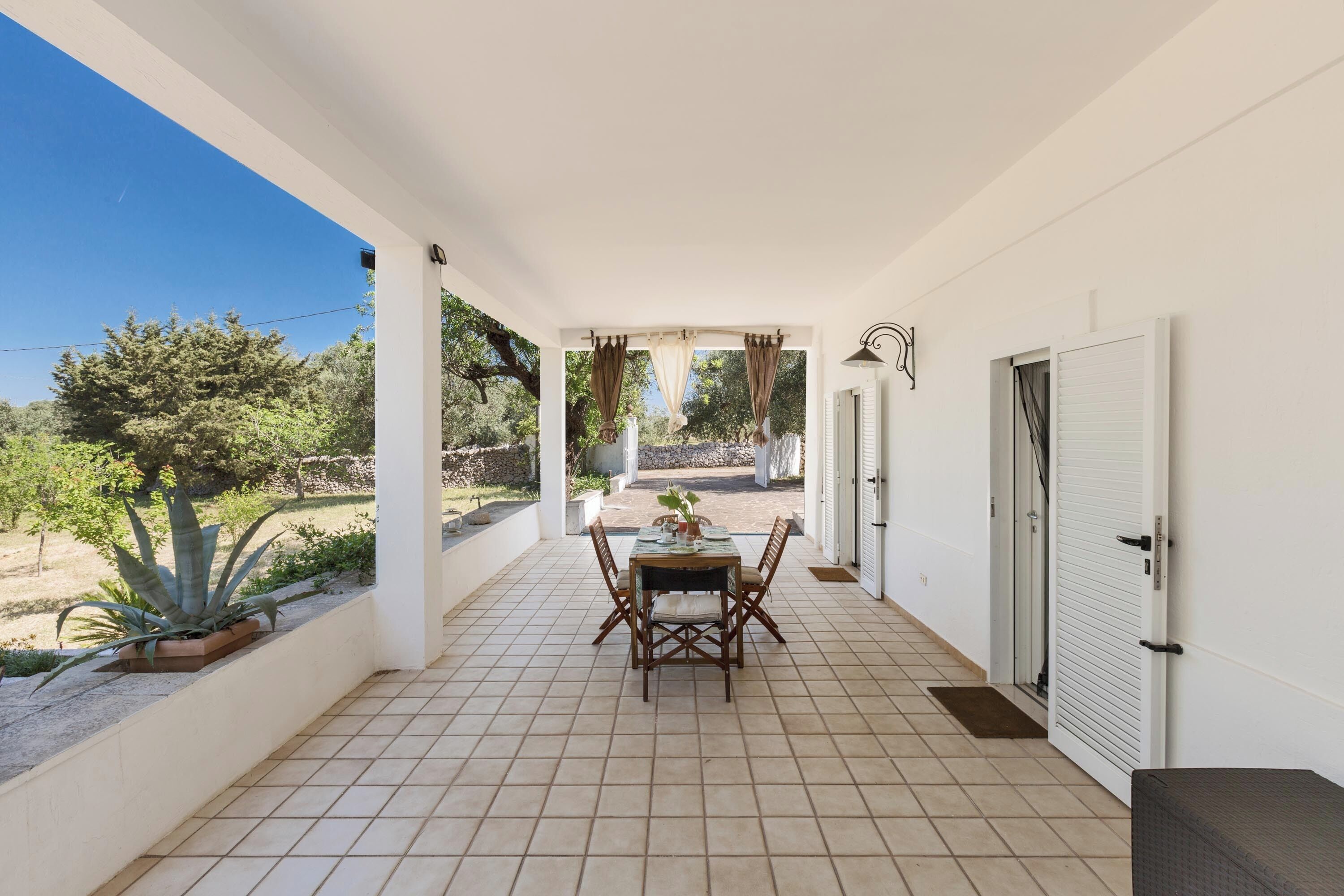 Une terrasse couverte avec un sol carrelé, une table et des chaises, et une vue sur un jardin et des arbres.