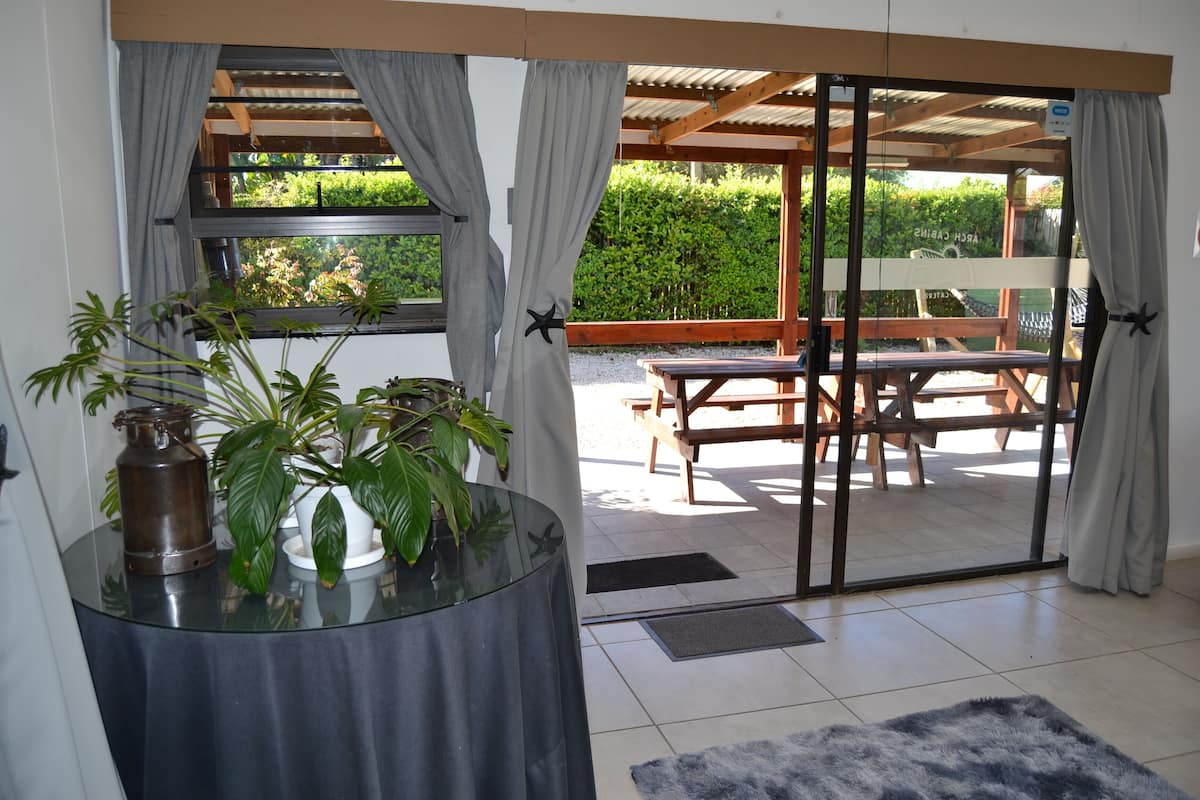 Quarto com mesa de vidro, uma planta em vaso e vista para um pátio externo com banco e mesa de madeira.