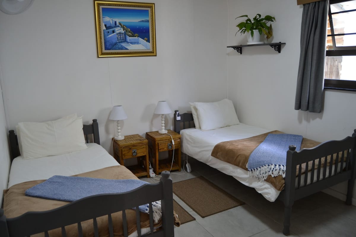 Quarto de hotel com duas camas, mesa de cabeceira com luminárias, prateleira pequena com uma planta e janela com cortinas.