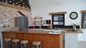 A modern kitchen with a wooden bar area, bar stools, a refrigerator, and a clock on the wall. - Arch Cabins Self Catering (Storms River)