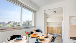 A modern dining area with a large window, a wooden table, and a view of the cityscape. - l'Emérillon - Appt à 200m de la plage (Saint-Cast-le-Guildo)