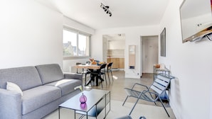 A modern living room with a grey sofa, a coffee table, and a dining area with a wooden table and chairs. There is a kitchen visible in the background. - l'Emérillon - Appt à 200m de la plage (Saint-Cast-le-Guildo)