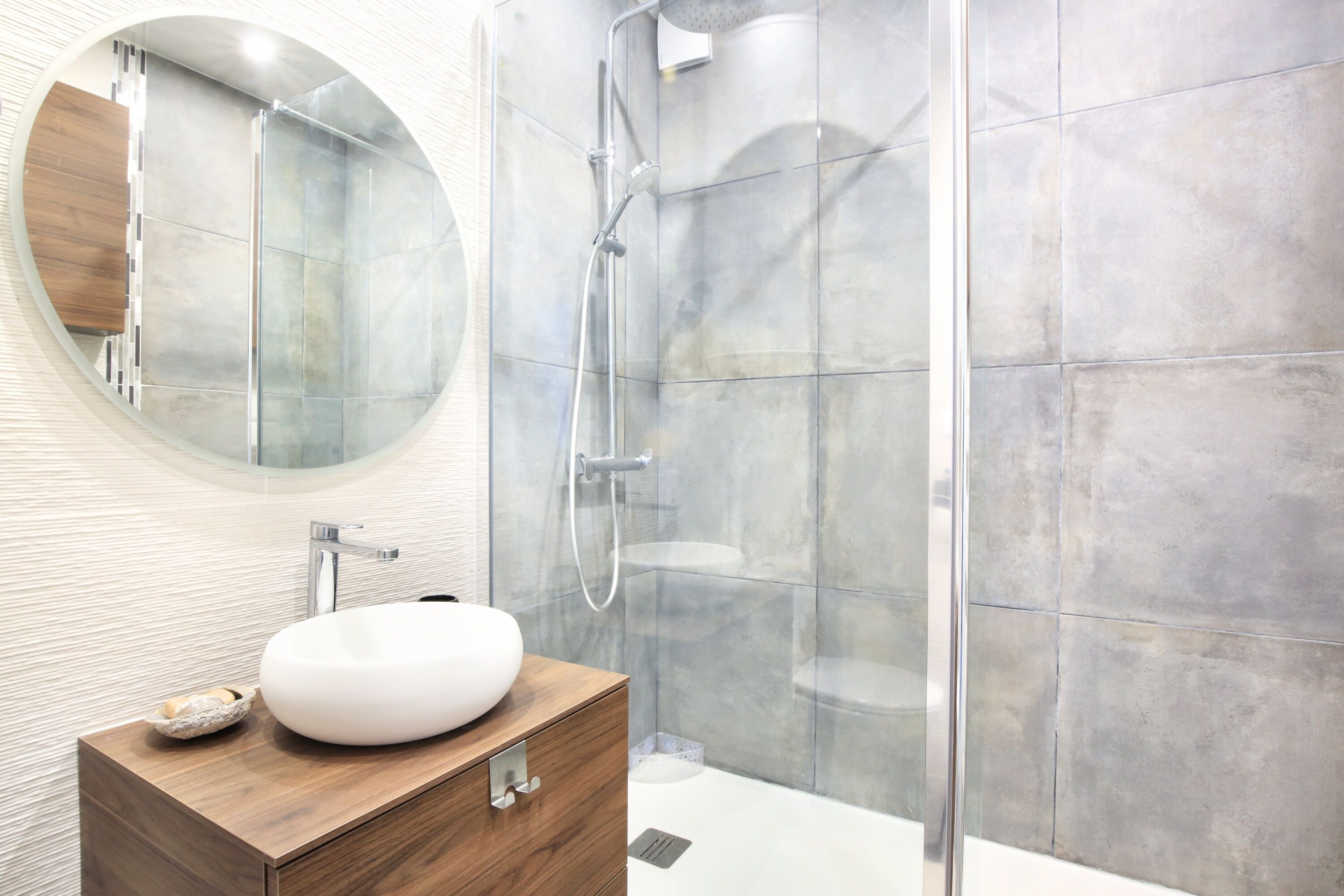 Een moderne badkamer met een ronde spiegel, een houten wastafelmeubel, een witte wasbak en een douche met glazen wanden.