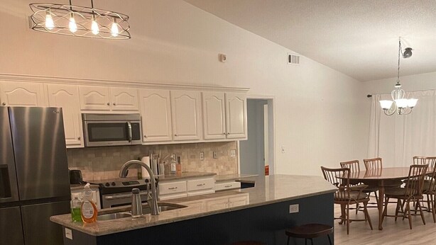 Una cocina moderna con armarios blancos, electrodomésticos de acero inoxidable, una isla central con taburetes y un comedor con sillas de madera.