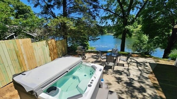 Un jacuzzi en un patio trasero con cerca de madera, una mesa, sillas y vista a un lago.