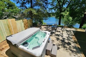 A hot tub in a backyard with a wooden fence, a table, chairs, and a view of a lake. - Little Blue Lake Front Cottage + Hot Tub (Greenville)
