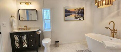 A bathroom with a white toilet, a black vanity with gold fixtures, a white bathtub, and a large window with a view of trees.